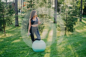 Outdoor shot of sporty young woman holds bottle of fresh water, listens music in earphones, holds modern smartphone, leans knee on