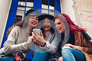 Outdoor shot of three young women looking at smartphone and laughing by cafe. Girls talking and having fun
