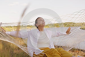Outdoor shot of young adult Caucasian female wearing white shirt sitting on hammock on the bank of the river, looking up at sky