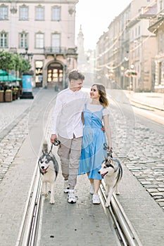 Outdoor shot of young couple man and woman, walking with two grey husky dogs, hugging each other on old European city