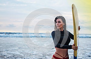 Outdoor shot of young girl waxes surfboard for safe riding waves, dressed in black swimsuit, takes care of safety, with white