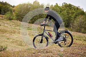 Outdoor shotof man riding mountain bike in meadow, sporty male dresses black track suit and cap, spending free time in active way