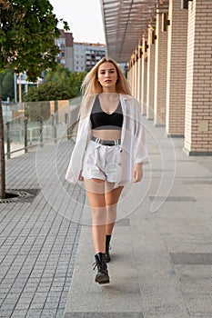 Outdoor street style photo, beautiful blonde girl in white shorts and black boots walks along modern building