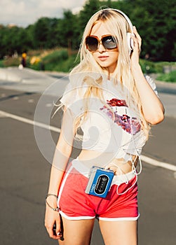 Outdoor summer fashion stylish portrait of young pretty blonde girl posing in vinage sunglasses and listening to music with h