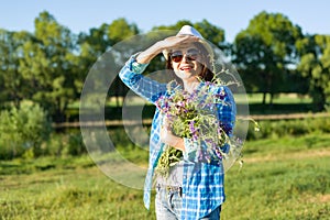 Outdoor summer portrait of an adult woman with bouquet of wildflowers, straw hat and sunglasses. Nature background, rural landscap