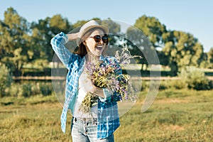 Outdoor summer portrait of adult woman with bouquet of wildflowers, straw hat and sunglasses. Nature background, rural landscape,