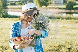 Outdoor summer portrait of adult woman with strawberries, bouquet of wildflowers, straw hat and sunglasses. Nature background,