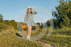 Outdoor summer portrait of girl teenager walking in rural country road in dress and hat with basket of strawberries. Nature