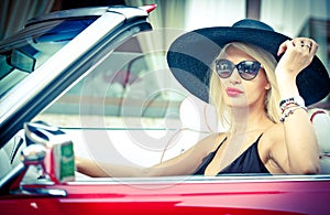 Outdoor summer portrait of stylish blonde vintage woman driving a convertible red retro car. Fashionable attractive fair hair girl