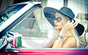 Outdoor summer portrait of stylish blonde vintage woman driving a convertible red retro car. Fashionable attractive fair hair girl