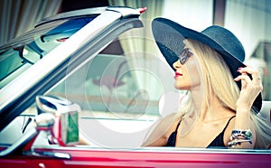 Outdoor summer portrait of stylish blonde vintage woman driving a convertible red retro car. Fashionable attractive fair hair girl