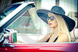 Outdoor summer portrait of stylish blonde vintage woman driving a convertible red retro car. Fashionable attractive fair hair girl