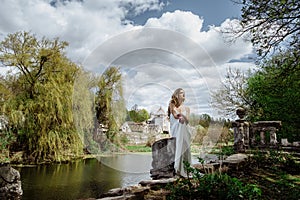 Outdoor summer portrait of young pretty cute girl. Beautiful woman posing at old bridge. in white dess siting near stone railing.