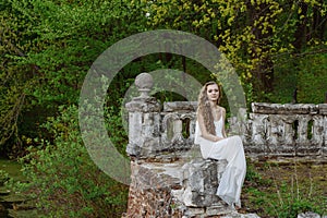 Outdoor summer portrait of young pretty cute girl. Beautiful woman posing at old bridge. in white dess siting near stone railing.