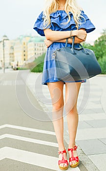 Outdoor summer smiling lifestyle portrait of pretty young woman with big blue handbag and hi heels shoes. Long blond hairs, blue