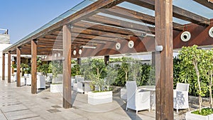 On the outdoor terrace, under a wooden canopy with a glass roof, there are white wicker chairs and tables
