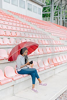 Outdoor view of the afro-american woman in the earphones, charring via the mobile phone while sitting on the stadium.