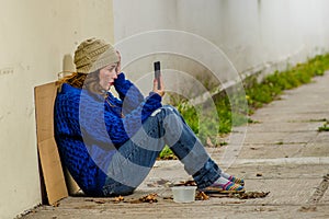 Outdoor view of homeless woman begging on the street in cold autumn weather sitting on the floor at sidewalk with a