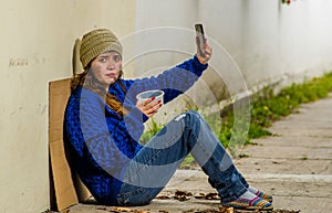 Outdoor view of homeless woman begging on the street in cold autumn weather sitting on the floor at sidewalk taking a