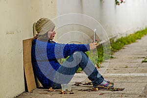 Outdoor view of homeless woman begging on the street in cold autumn weather sitting on the floor at sidewalk taking a