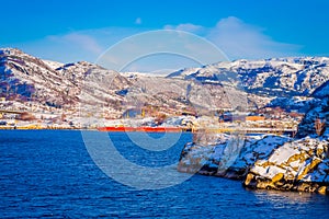 Outdoor view of huge red boat in a shrore with wooden houses a long in the coast from Hurtigruten voyage, Northern