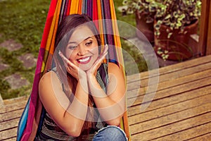 Outdoor view of smiling young beautiful woman relaxing in a hammock with both hands under her chin, in wooden nature