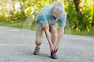 Outdoor view of sporty healthy male pensioner stretches leg, pulled muscle, dressed in sportswear and sneakers, stands on asphalt,
