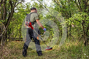 Outdoor view of worker wearing protective clothing and gloves while using a lawn trimmer mower cutting grass in a nature