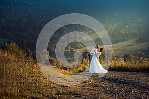 Outdoor wedding portrait of the adorable cheerful young newlywed couple softly hugging on the road in the countryside