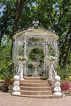 Outdoor white metal gazebo with roof in a garden against green trees
