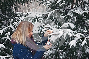 Outdoor winter Christmas portrait of Woman in snowy forest. Smiling Caucasian blonde woman decorates fir tree for the holiday