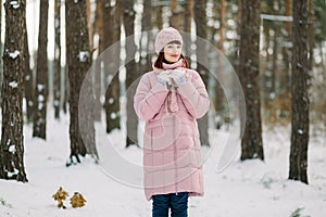 Outdoor winter portrait of pretty young woman wearing pink coat and stylish knitted hat and scarf, standing in beautiful