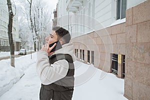 Outdoor winter portrait for young handsome man with the phone. Beautiful teenager in his jacket and vest posing on a city street,