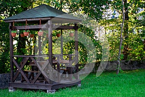 Outdoor wooden gazebo over summer landscape background. Wood brown arbour on green lawn. Summertime lush foliage pine trees blue