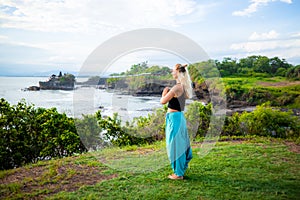 Outdoor yoga practice. Young woman standing on the grass. Hands in namaste mudra, closed eyes. Tanah Lot temple, Bali