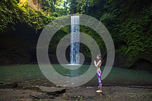 Outdoor yoga practice. Young woman standing near waterfall. Hands in namaste mudra, closed eyes. Preparing for Sun salutation