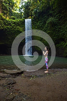 Outdoor yoga practice. Young woman standing near waterfall. Hands in namaste mudra, closed eyes. Preparing for Sun salutation