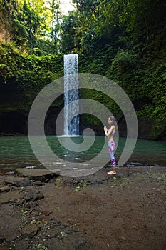 Outdoor yoga practice. Young woman standing near waterfall. Hands in namaste mudra, closed eyes. Preparing for Sun salutation