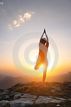 Outdoor yoga silhouette, young woman in tree pose. A woman practices yoga on the green grass on top of a mountain with a