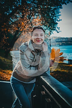 Outdoors fashion portrait of happy young woman walking in autumn park. Model girl with a bright orange autumn leaf in hand leans