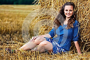 Outdoors fashion portrait of young woman in blue dress sitting near haystack