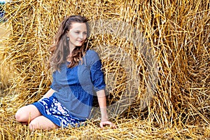 Outdoors fashion portrait of young woman in blue dress sitting near haystack