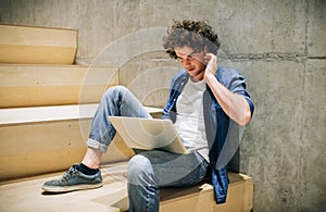 Outdoors image of young hipster male with curly hair using laptop for chatting online with friends, connected to free wireless on