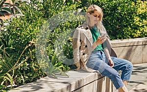 Outdoors imaget of young woman writing messages on smartphone, sitting outdoor in the city at sunny day.