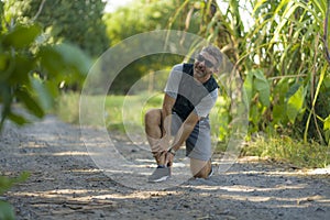 Outdoors jogging accident - young attractive runner man in sport suffering ankle problem during running workout in city park