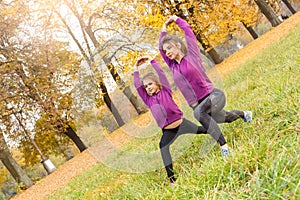Outdoors leisure. Sisters doing lunge exercise hands up in the autumn park looking aside joyful