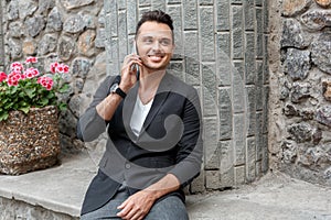 Outdoors leisure. Young man in suit on city street sitting talking on smartphone smiling joyful