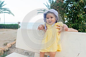 outdoors lifestyle portrait of adorable and happy baby girl in cute hat hold by her mothers hands on park bench looking excited