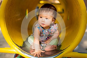 outdoors lifestyle portrait of beautiful and adorable 9 month old baby girl sitting on yellow tube at children playground in city