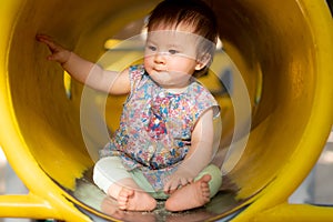 outdoors lifestyle portrait of beautiful and adorable 9 month old baby girl sitting on yellow tube at children playground in city